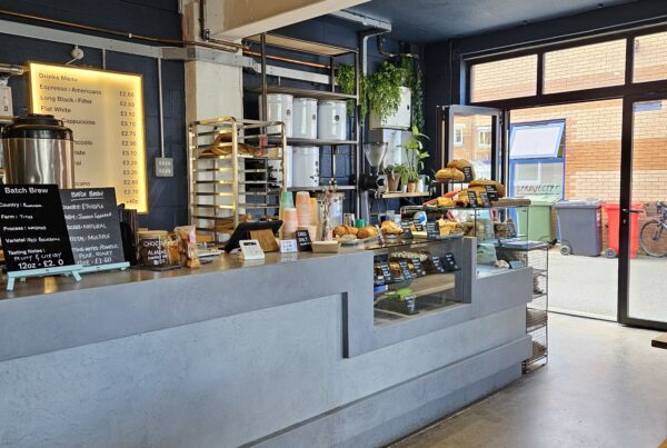 Interior of an Missing Bean coffee shop with a grey concrete counter, a backlit drinks menu and batch brew tasting boards, a display of pastries and sourdough loaves, and open glass doors letting in daylight.