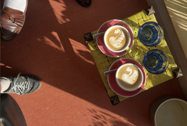 Two coffees with swan latte art on red saucers beside two blue glasses of water on a yellow woven stool, with dappled sunlight and leaf shadows across a terracotta floor.