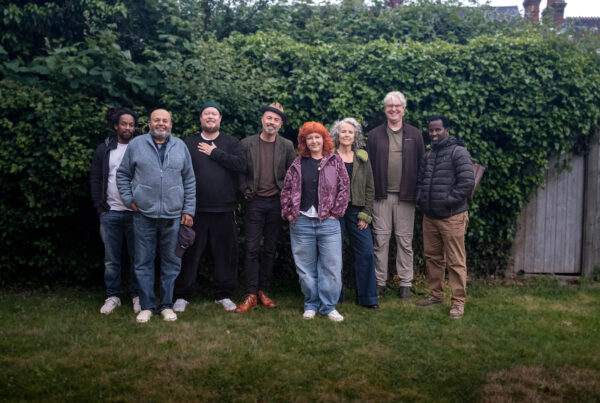 A group of eight people stand together on a lawn in front of a dense green hedge, smiling at the camera. The group is casually dressed in a mix of jackets, fleeces and jeans. One woman in the centre wears a purple patterned jacket and has bright red hair; another woman to her right has short silver hair and a green jacket. Two men wear wide-brimmed hats. The setting appears to be a garden in an overcast daylight.