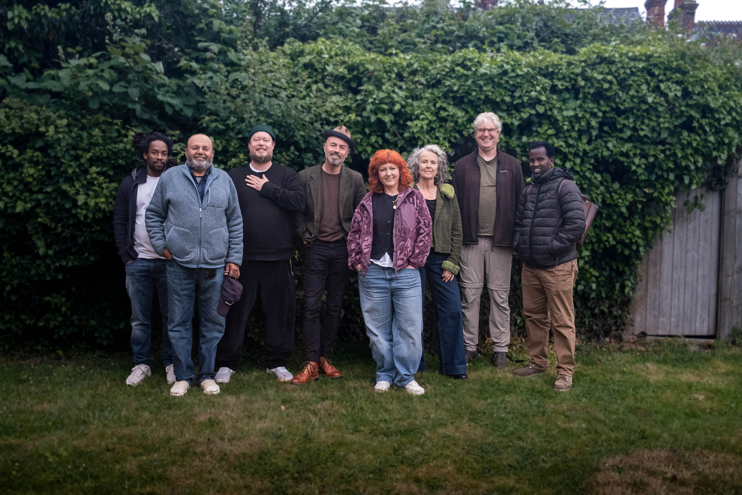 A group of eight people stand together on a lawn in front of a dense green hedge, smiling at the camera. The group is casually dressed in a mix of jackets, fleeces and jeans. One woman in the centre wears a purple patterned jacket and has bright red hair; another woman to her right has short silver hair and a green jacket. Two men wear wide-brimmed hats. The setting appears to be a garden in an overcast daylight.