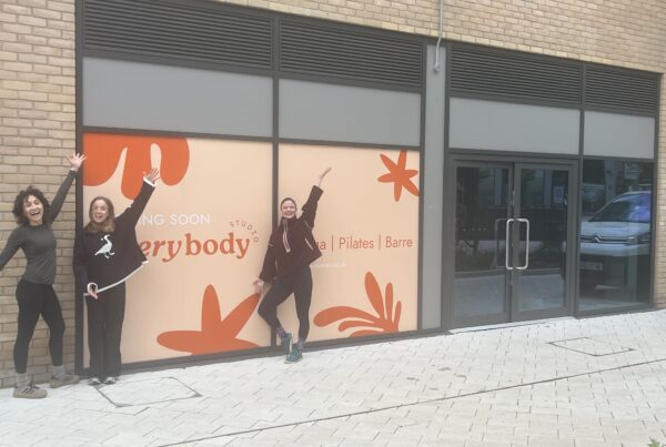 Three women celebrate outside the upcoming Everybody Studio, a yoga, Pilates and barre studio. The shopfront features large "Coming Soon" window graphics in a warm peach and burnt orange botanical design. All three women have their arms raised in excitement — one stands to the far left in a dark long-sleeved top and black leggings, one in the centre wears a black sweatshirt, and one strikes a dynamic pose in front of the signage in a dark zip-up jacket and patterned leggings. The entrance doors to the unit are visible to the right.