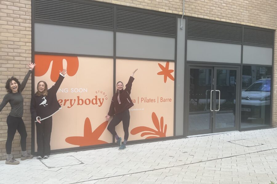 Three women celebrate outside the upcoming Everybody Studio, a yoga, Pilates and barre studio. The shopfront features large "Coming Soon" window graphics in a warm peach and burnt orange botanical design. All three women have their arms raised in excitement — one stands to the far left in a dark long-sleeved top and black leggings, one in the centre wears a black sweatshirt, and one strikes a dynamic pose in front of the signage in a dark zip-up jacket and patterned leggings. The entrance doors to the unit are visible to the right.