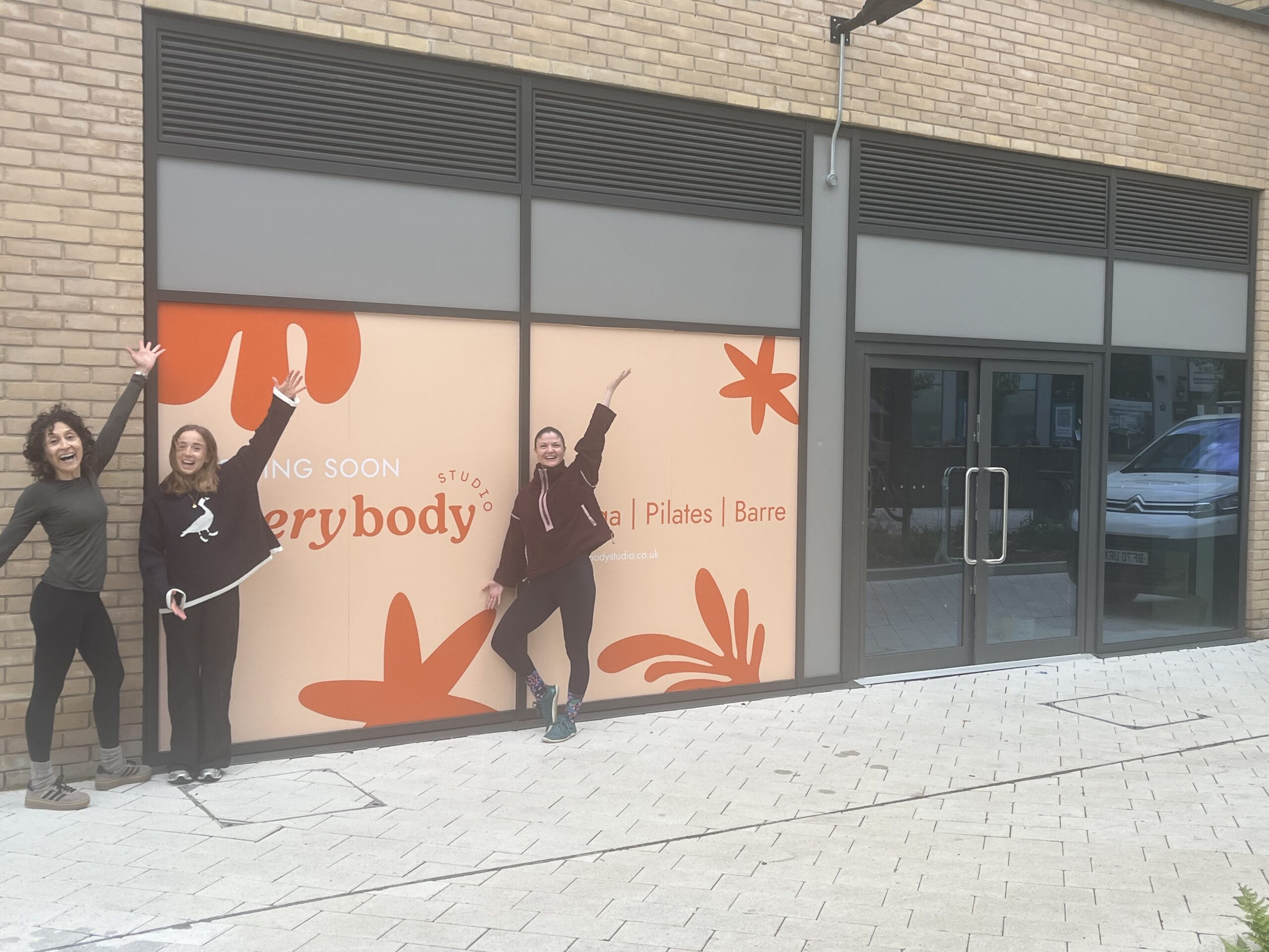 Three women celebrate outside the upcoming Everybody Studio, a yoga, Pilates and barre studio. The shopfront features large "Coming Soon" window graphics in a warm peach and burnt orange botanical design. All three women have their arms raised in excitement — one stands to the far left in a dark long-sleeved top and black leggings, one in the centre wears a black sweatshirt, and one strikes a dynamic pose in front of the signage in a dark zip-up jacket and patterned leggings. The entrance doors to the unit are visible to the right.